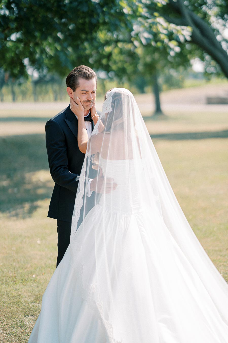 Bride and groom sharing an intimate moment outdoors, with the bride wearing a white gown and veil, gently touching the groom’s face, surrounded by lush greenery.