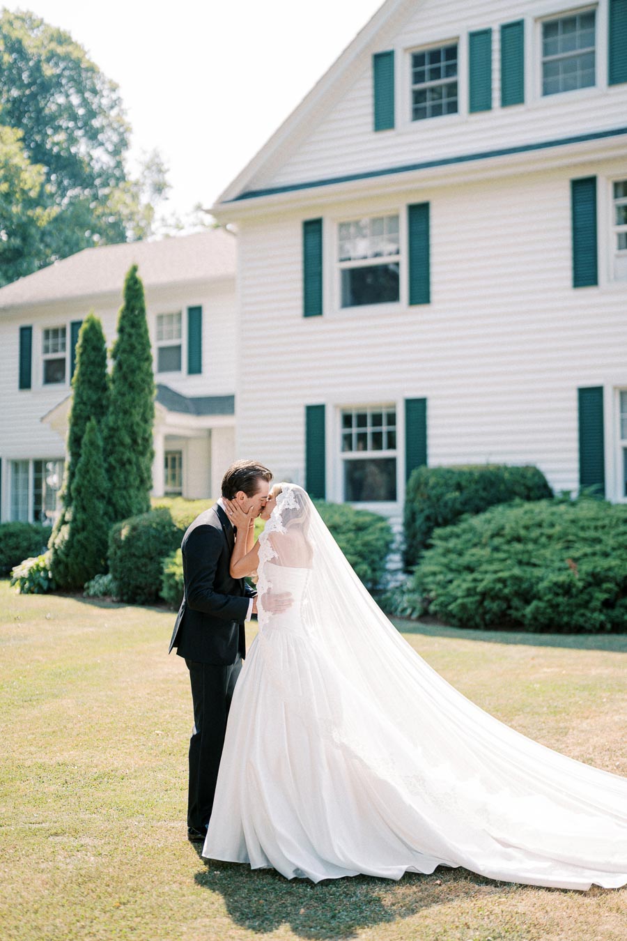 Bride and groom sharing a kiss in front of a white house with green shutters, bride wearing a long flowing white gown and veil, on a sunny day with lush greenery in the background.