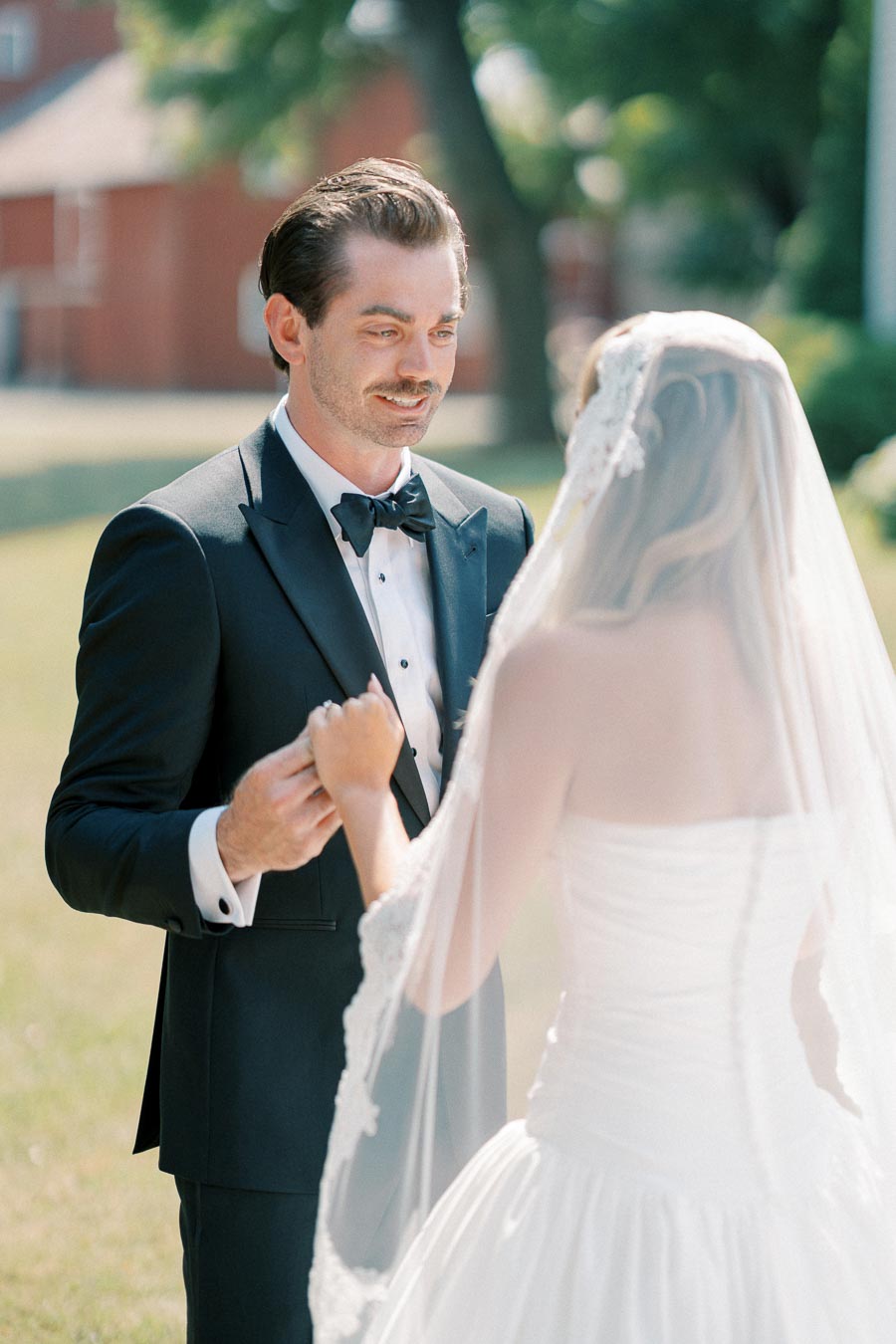A groom in a black tuxedo holds hands with a bride wearing a white wedding dress and veil, standing outdoors on a sunny day.