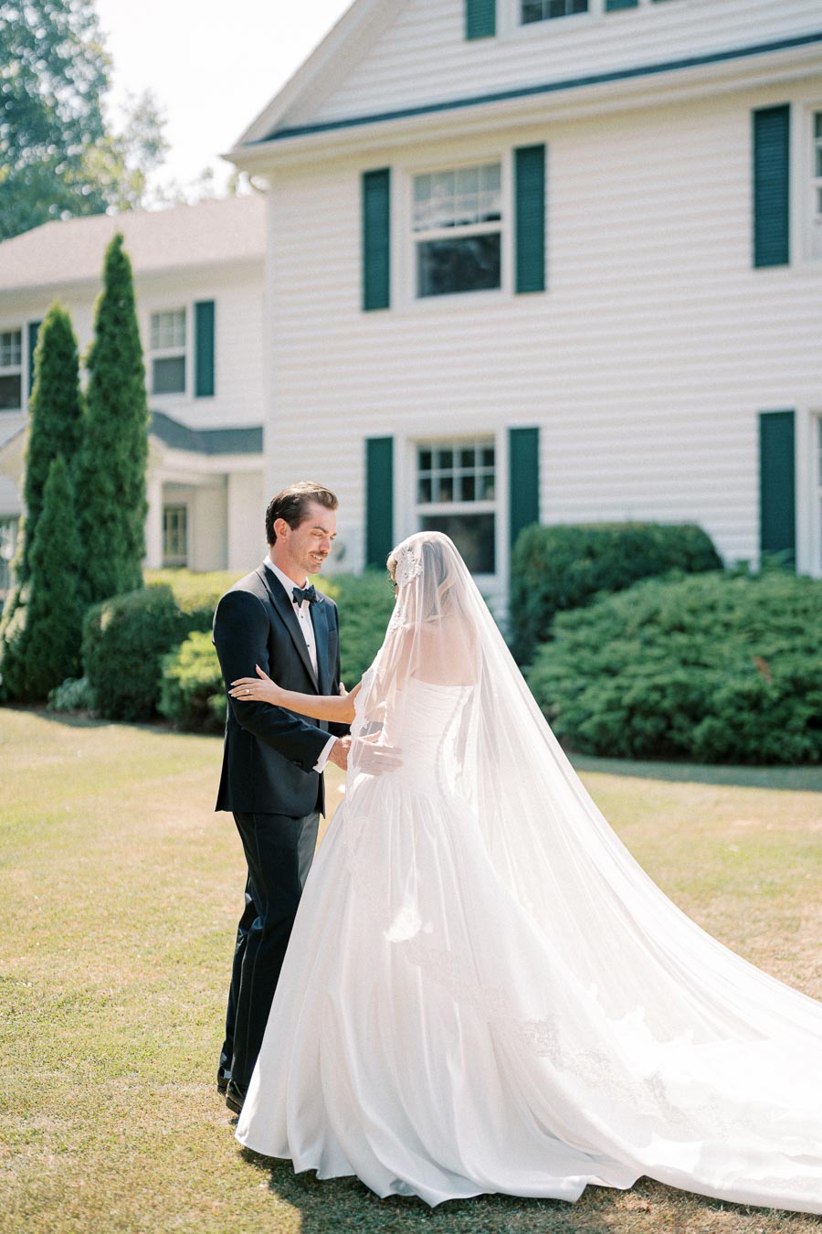 A bride and groom dressed in elegant wedding attire share a tender moment in front of a charming white house with green shutters and lush landscaping, capturing a beautiful outdoor wedding scene.