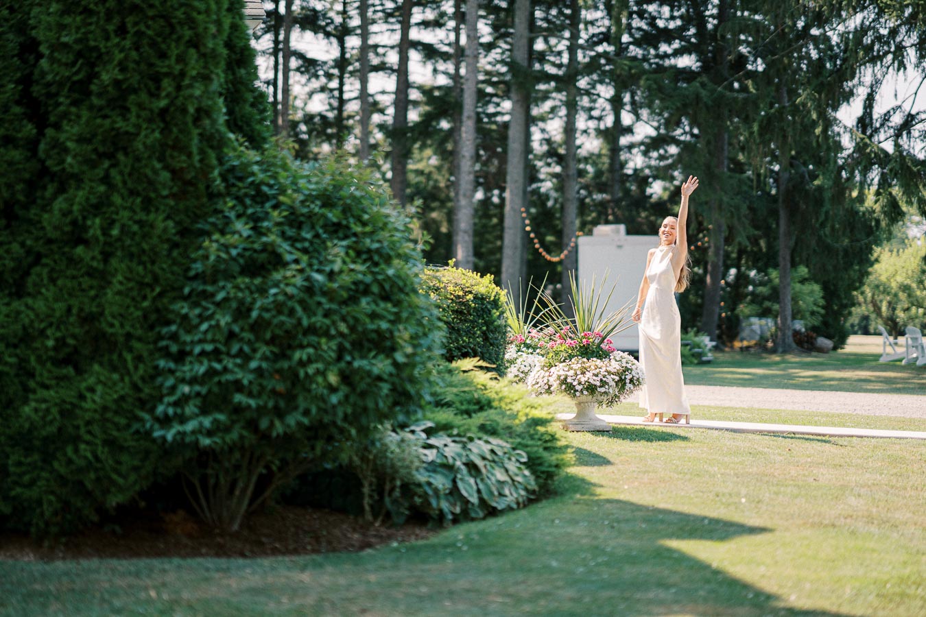 A woman in an elegant white dress stands joyfully in a lush garden setting, surrounded by vibrant greenery and colorful flowers, with tall trees in the background.