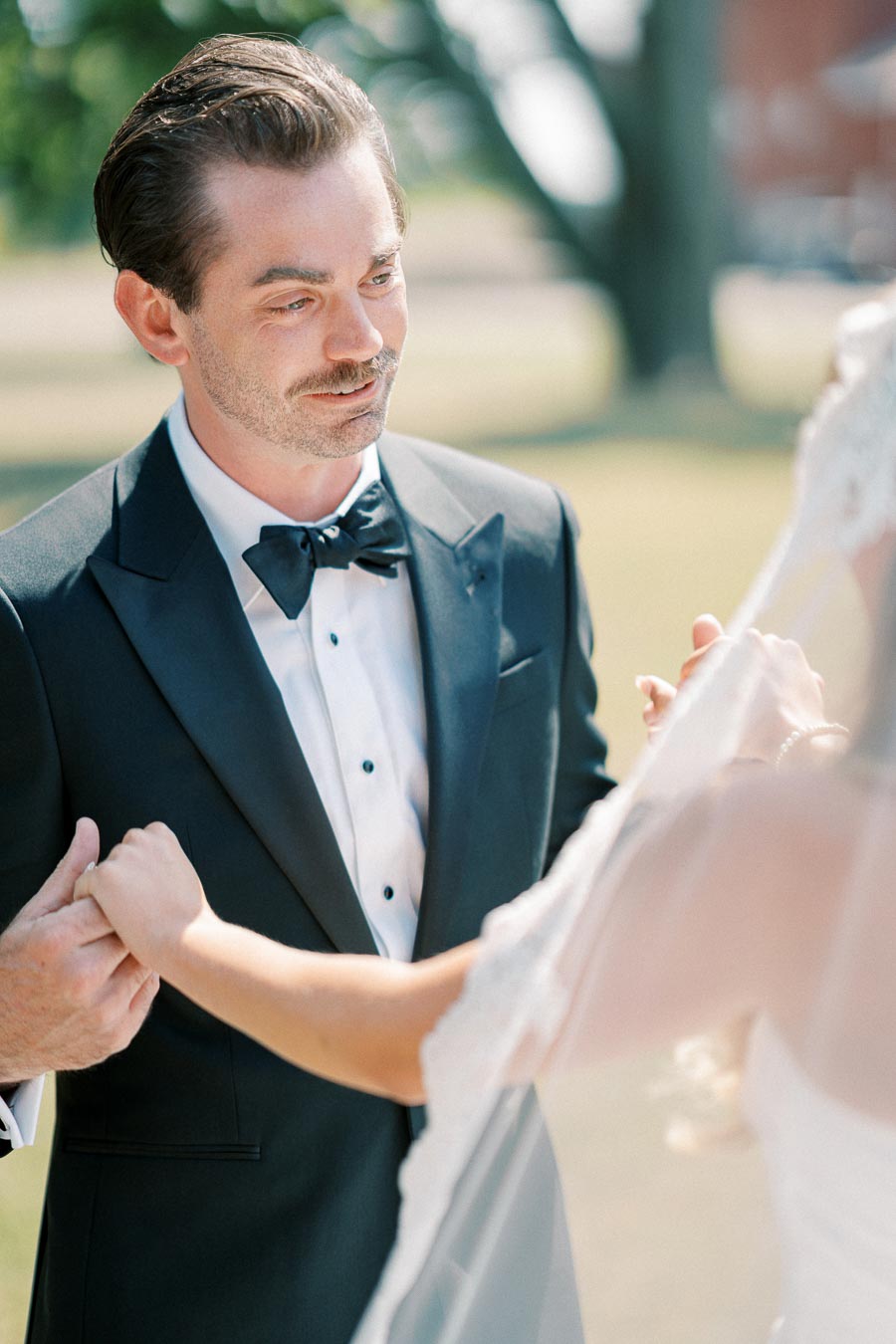 Groom in black tuxedo holding hands with bride in white dress, outdoor wedding ceremony setting