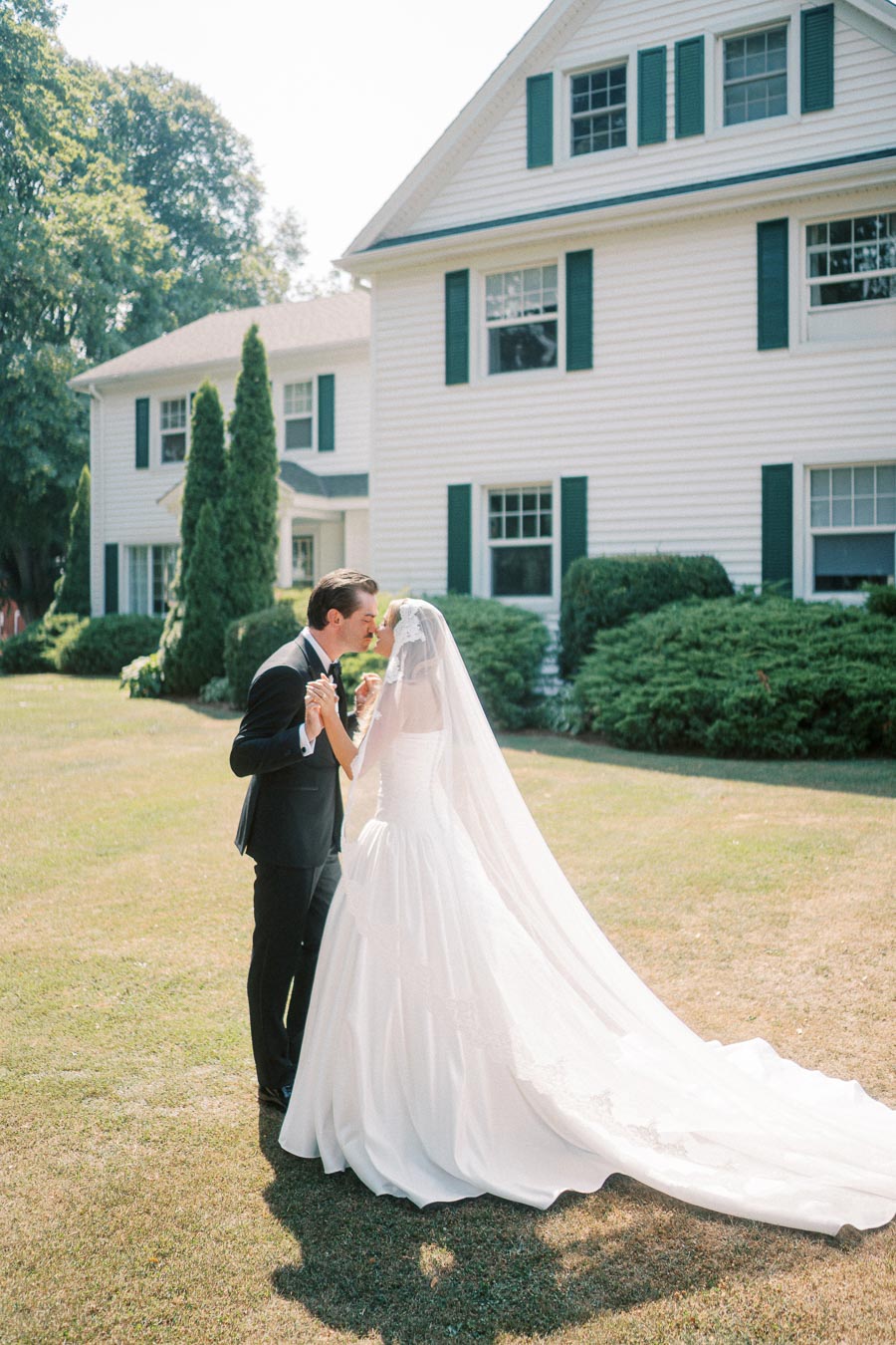 Bride and groom sharing a kiss in front of a white house with green shutters on a sunny day, showcasing a romantic outdoor wedding setting.