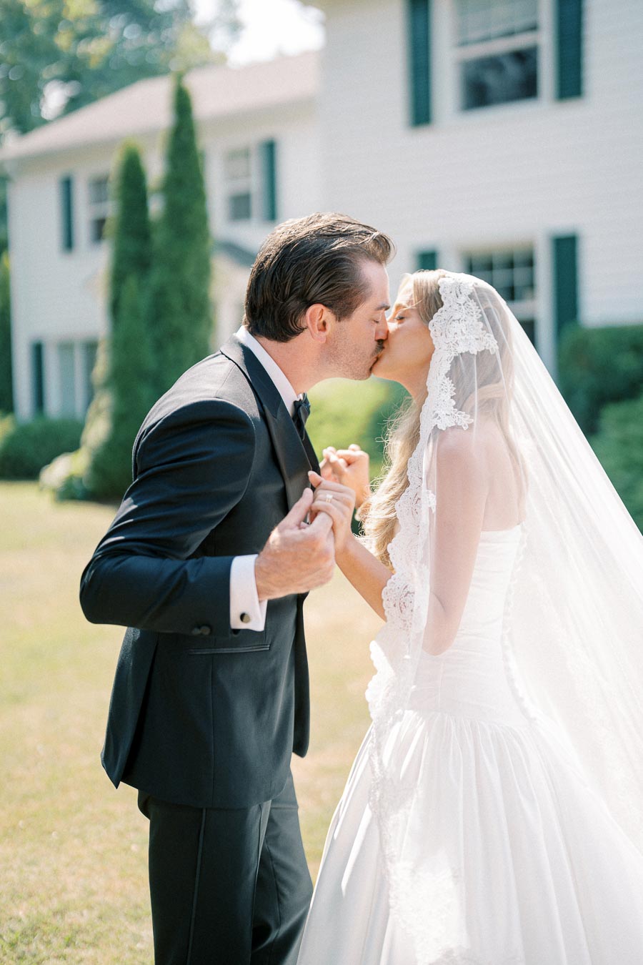 Bride and groom share a kiss outdoors in front of a white house, surrounded by greenery; the bride wears a lace veil and a white gown, while the groom is in a tailored black suit.