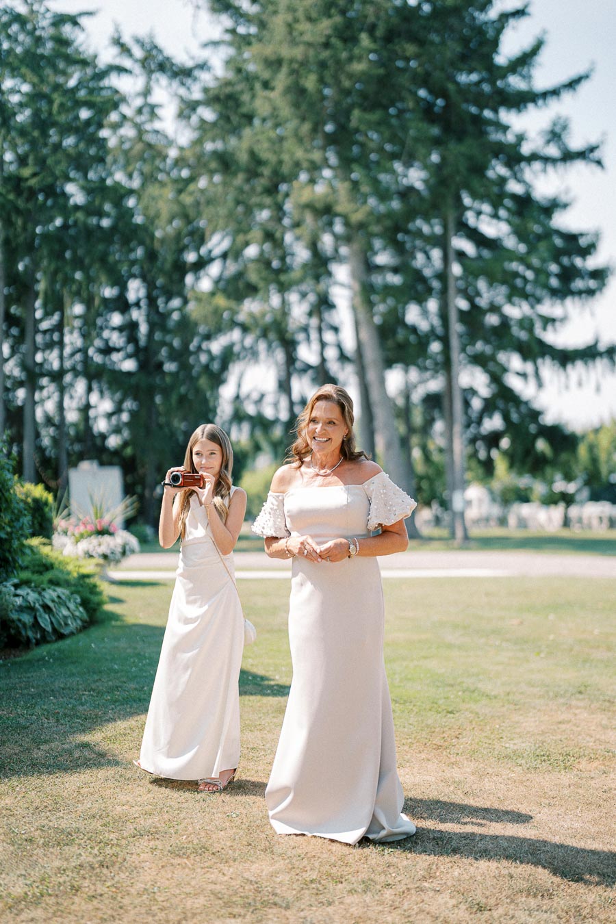 Two women in elegant white dresses enjoying an outdoor event, surrounded by lush greenery and tall trees on a sunny day.