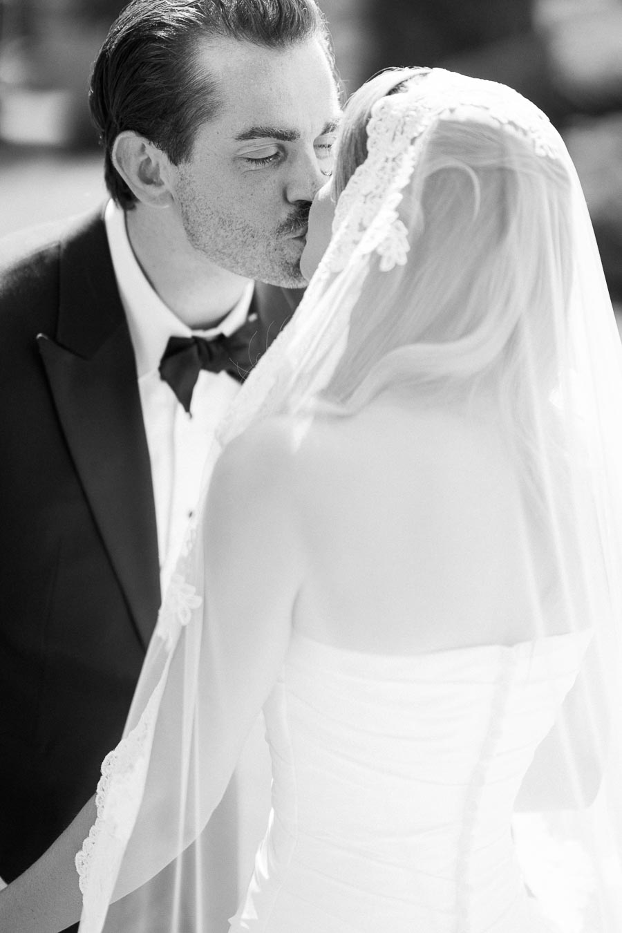 A black and white photo of a newlywed couple sharing a kiss on their wedding day. The groom is wearing a classic tuxedo, and the bride is in a lace-adorned veil and wedding dress, creating a romantic and timeless moment.