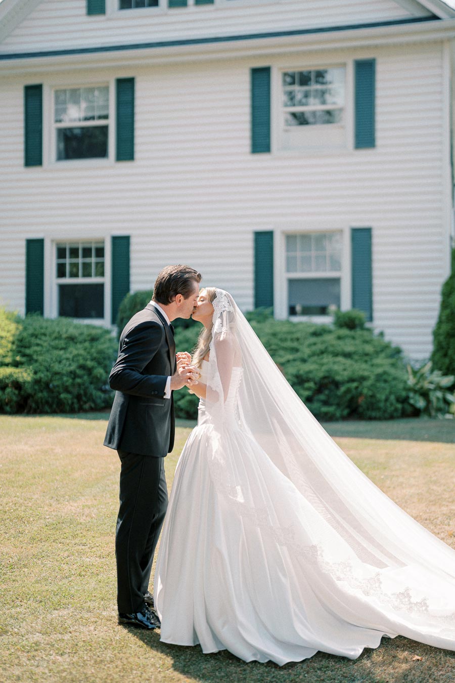 Elegant bride and groom share a romantic kiss in front of a classic white house, with the bride wearing a flowing lace wedding gown and veil, symbolizing timeless wedding romance.