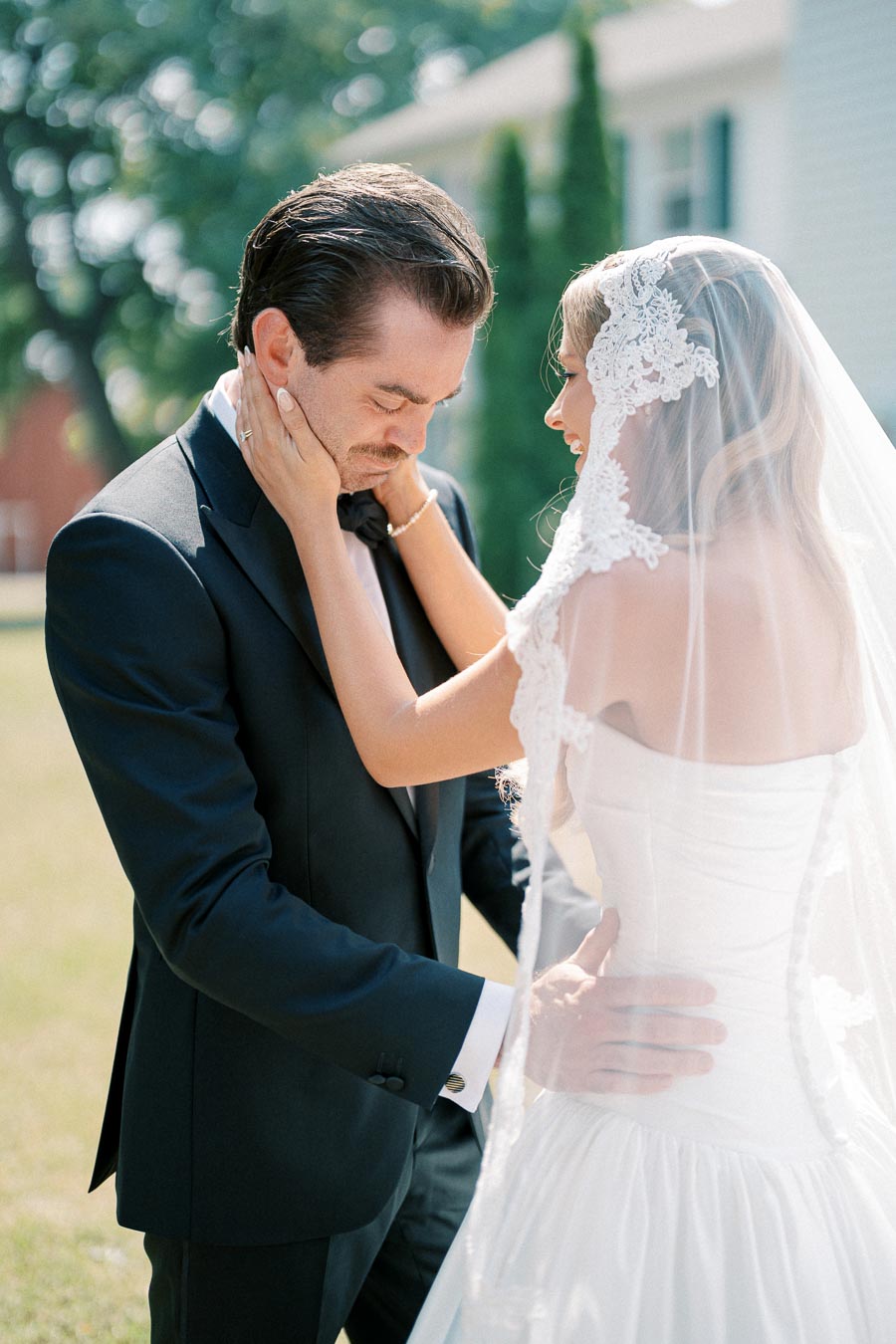 A bride and groom sharing a tender moment outdoors, with the bride touching the groom's face while wearing a lace veil and a white wedding dress, and the groom in a black suit, against a backdrop of greenery and a house.