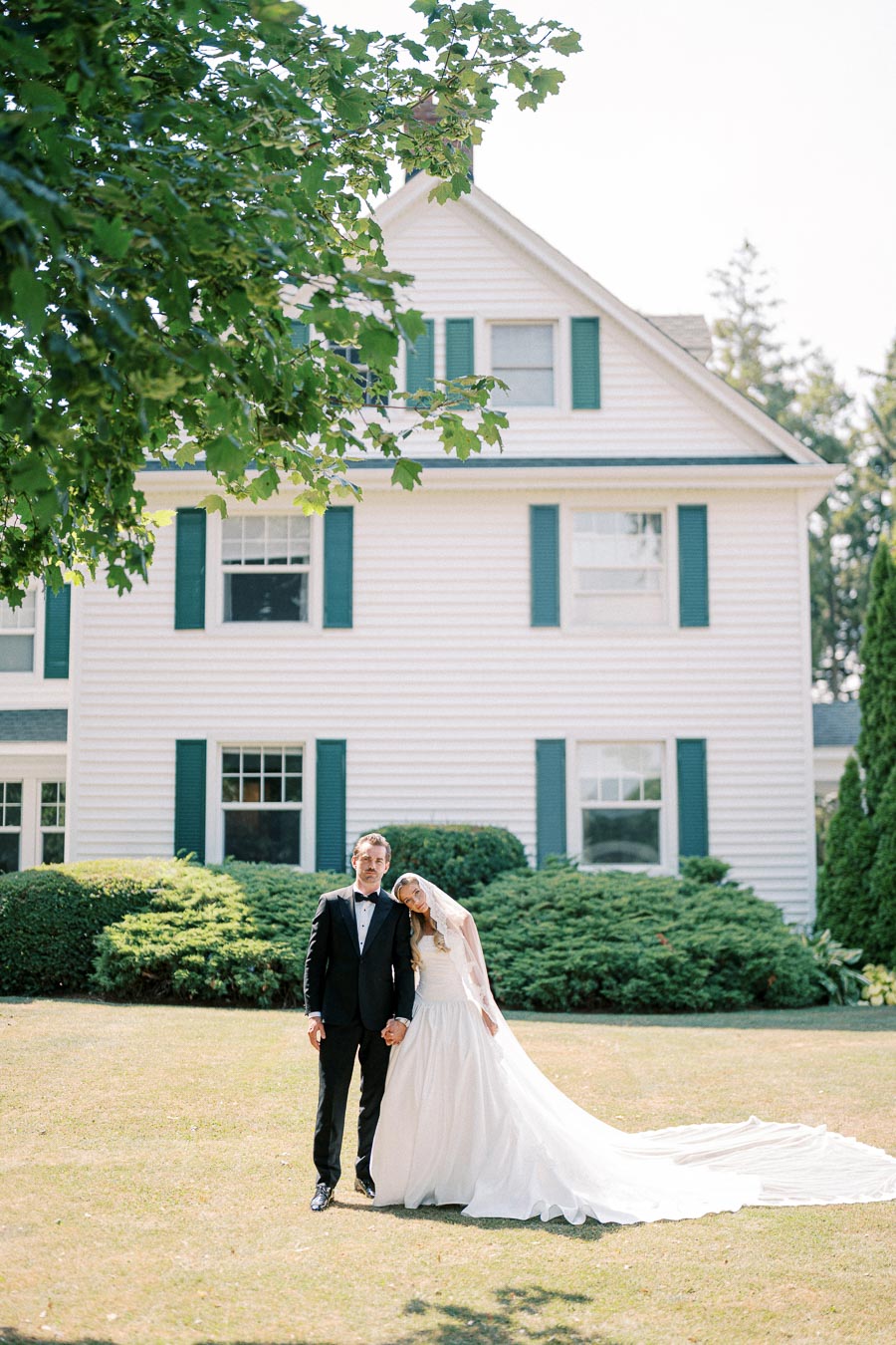 Elegant bride and groom posing on the lawn in front of a traditional white house with green shutters, showcasing a beautiful wedding scene under a vibrant tree.