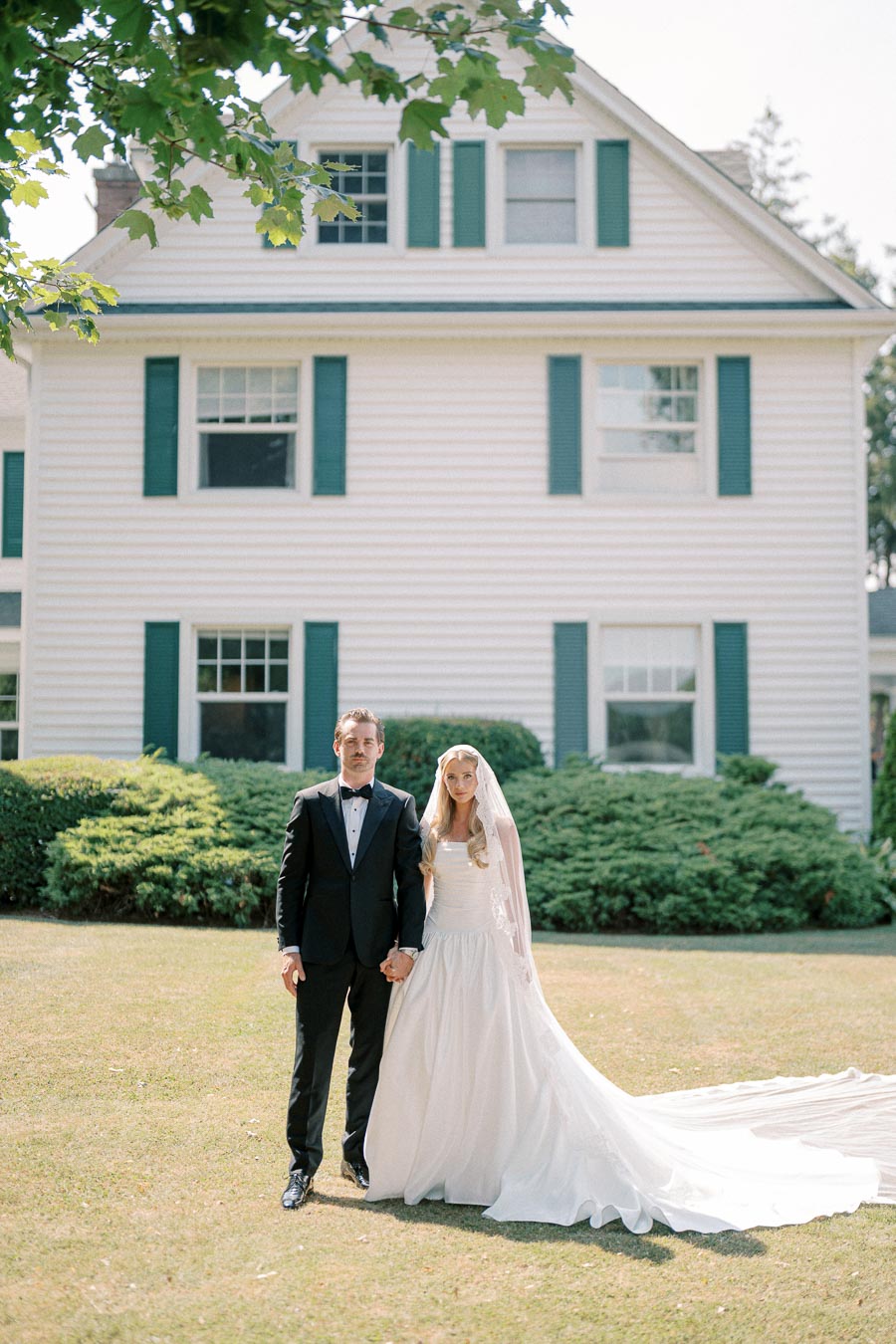 Elegant bride and groom pose in front of a classic white house with green shutters on a sunny day, showcasing a beautiful wedding dress with a long train and a sleek tuxedo.