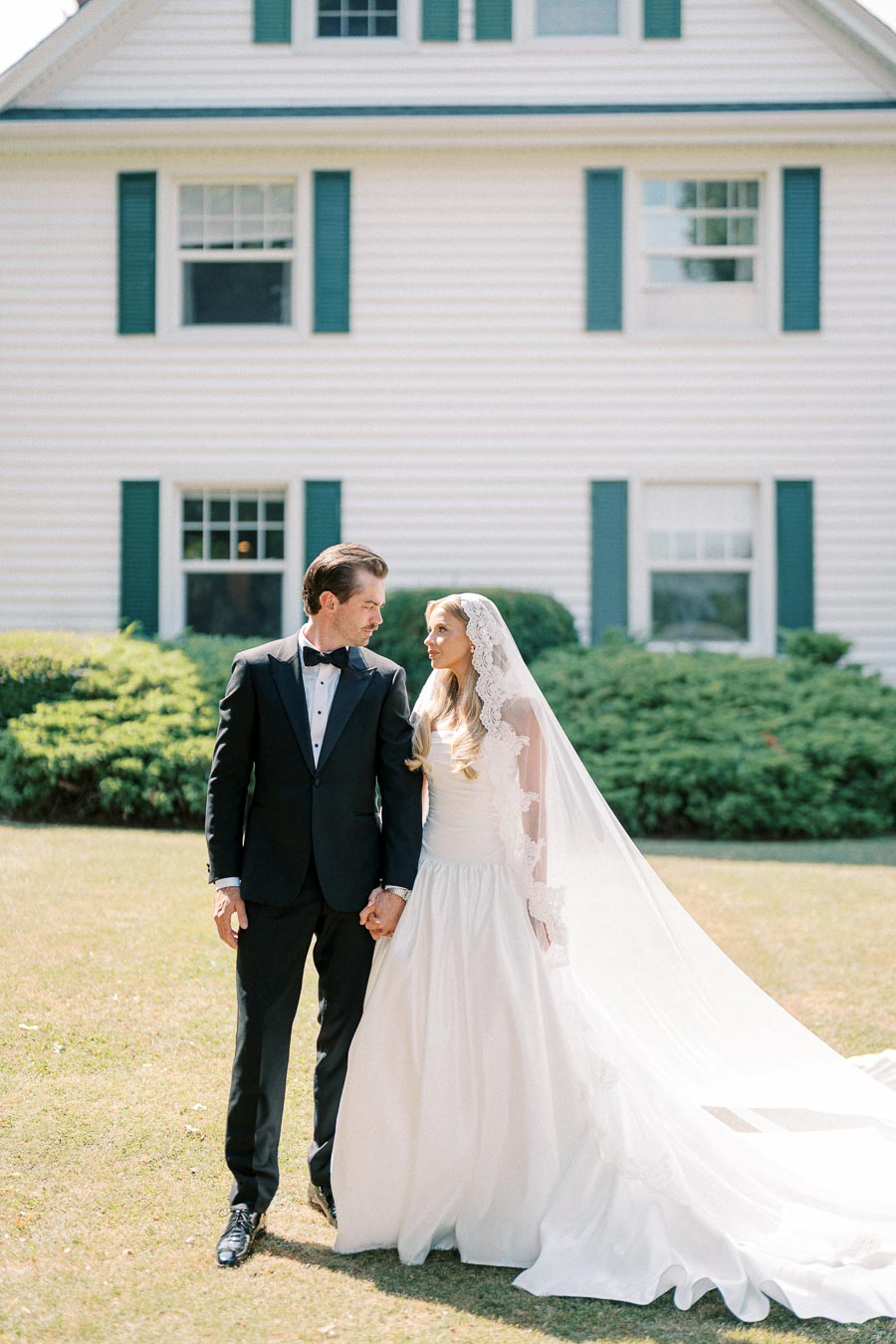 A bride in a flowing white gown and lace veil stands beside a groom in a classic black tuxedo, both holding hands and gazing at each other in front of a charming white house with green shutters.