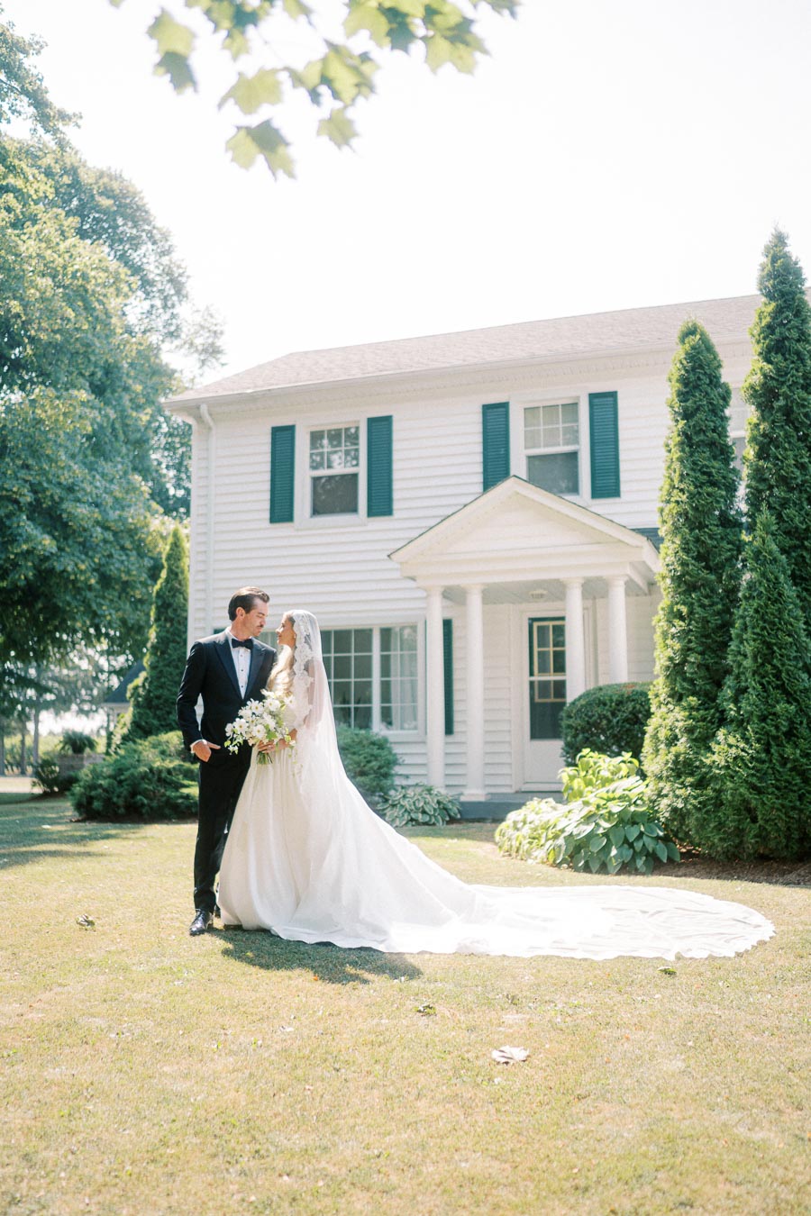 Elegant bride and groom in wedding attire standing on a lush green lawn in front of a classic white house, surrounded by trees, on a sunny day; wedding photography.