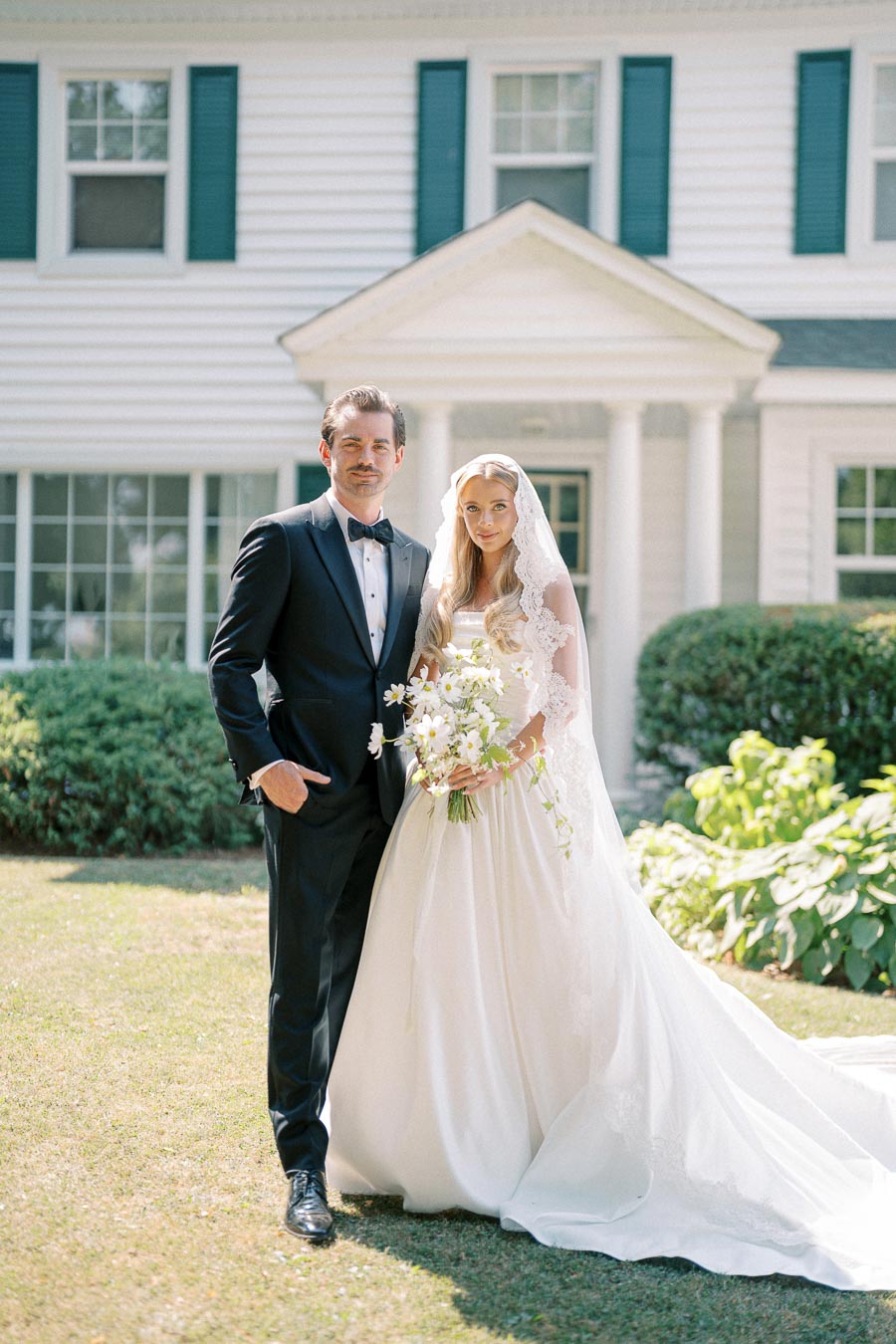 Elegant bride and groom posing outside a classic white house, with the bride in a lace wedding gown holding a bouquet and the groom in a formal black tuxedo, both smiling under a sunny sky.