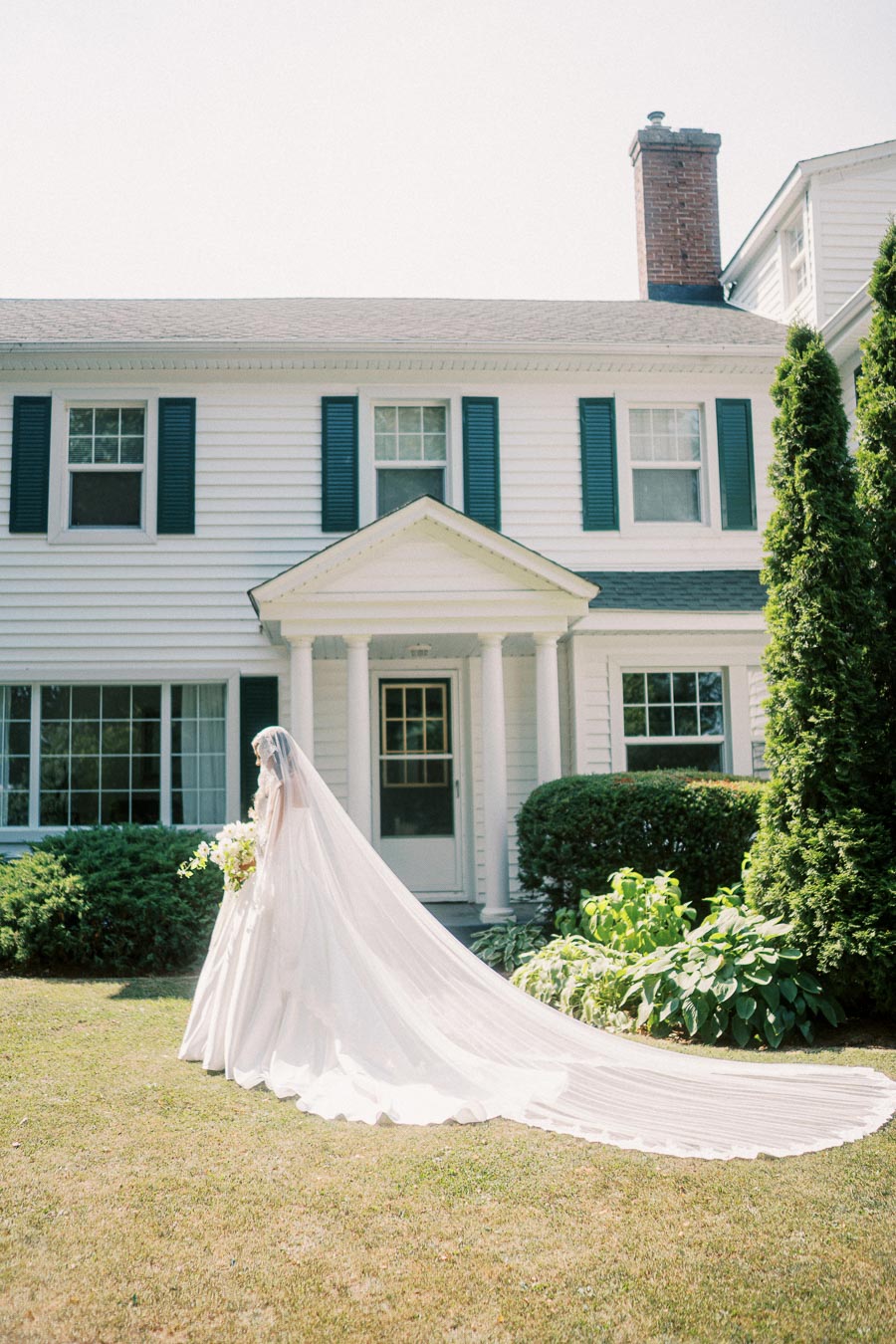 A bride in a flowing white wedding dress and veil stands in front of a classic white house with dark shutters, surrounded by greenery, on a sunny day.