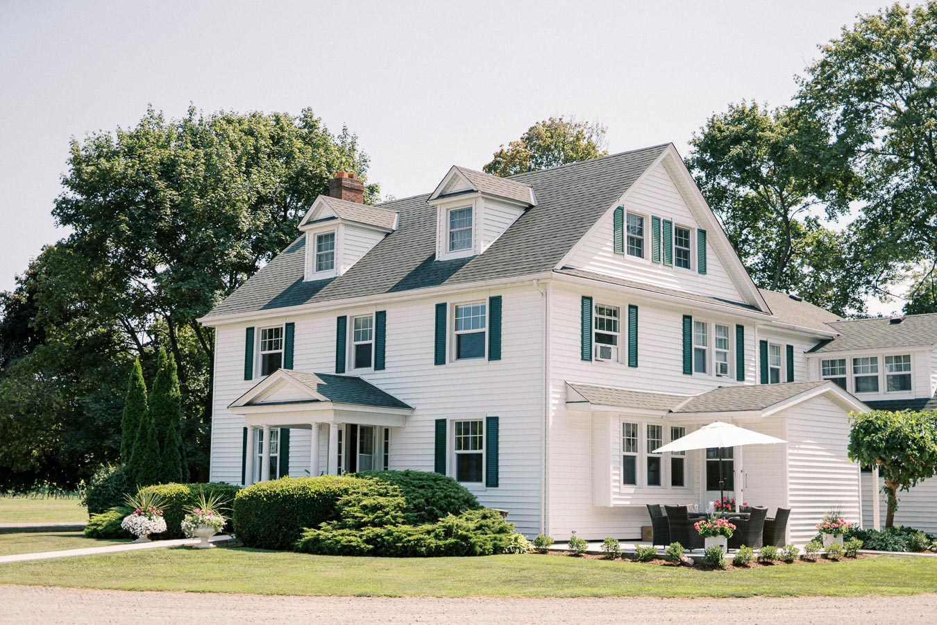 Colonial-style white house with green shutters, featuring a neatly landscaped front yard and patio set under an umbrella, surrounded by lush trees on a sunny day.