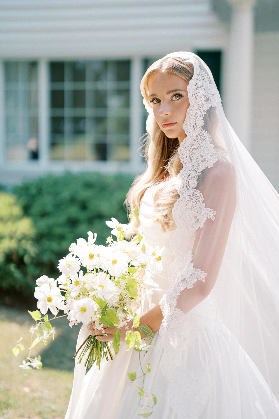 A bride in a white lace veil holding a bouquet of white flowers outdoors, with a windowed building in the background.