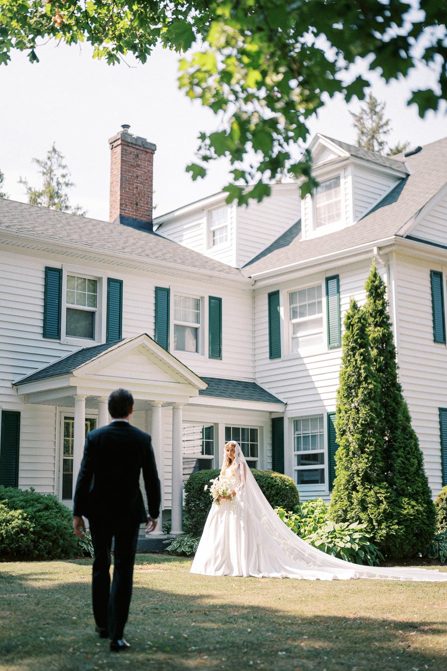 A bride in an elegant white gown holding a bouquet walks towards a man in a suit, set against the backdrop of a charming white house with green shutters and lush greenery on a sunny day.