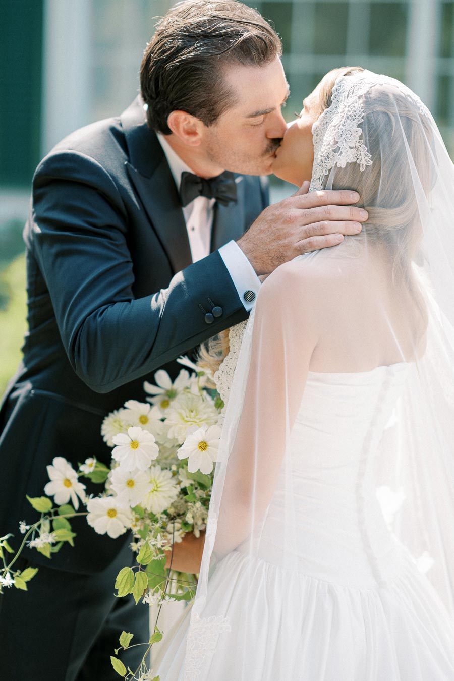 A bride in a lace veil and groom in a tuxedo share a romantic kiss, with the bride holding a bouquet of white flowers.