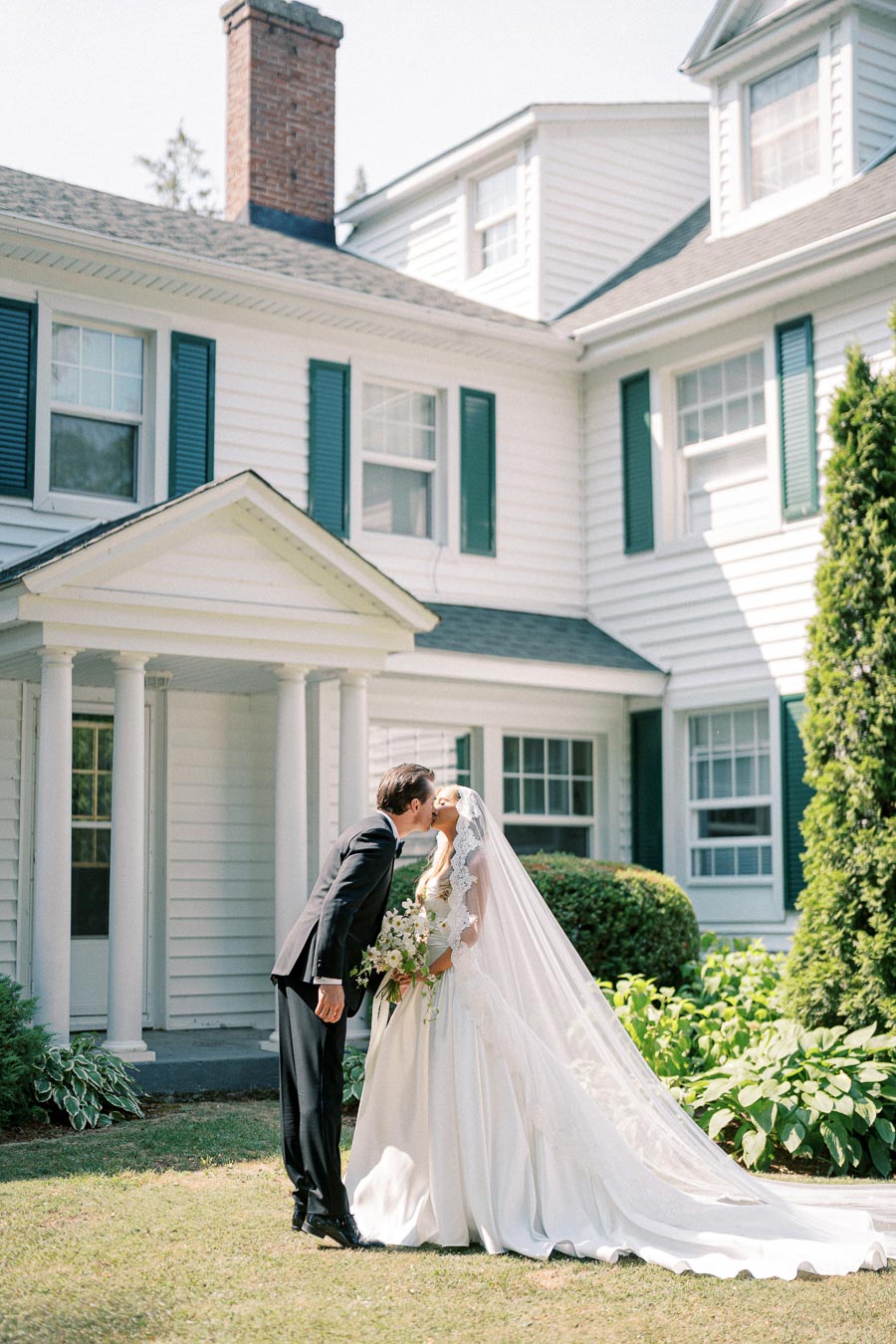 A bride and groom share a kiss in front of a classic white colonial-style house with green shutters, surrounded by lush greenery on a sunny day.