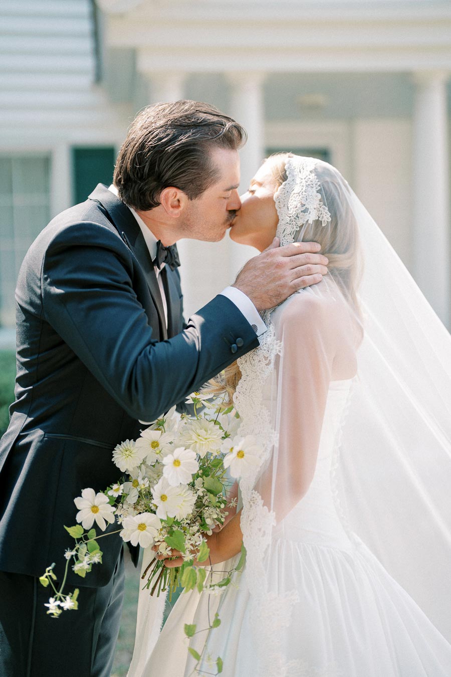 Elegant bride and groom share a romantic kiss outdoors, the bride holding a bouquet of white flowers, showcasing a lace wedding dress and veil, while the groom wears a classic black tuxedo.