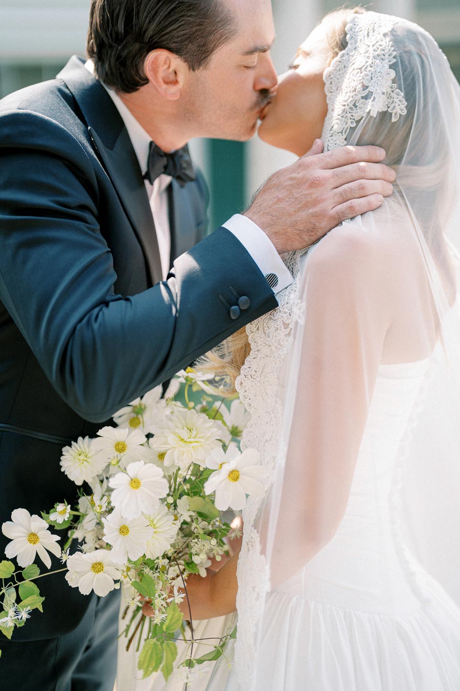 A bride and groom share a romantic kiss on their wedding day, with the bride holding a bouquet of white flowers and wearing a lace veil, while the groom is dressed in a formal suit.