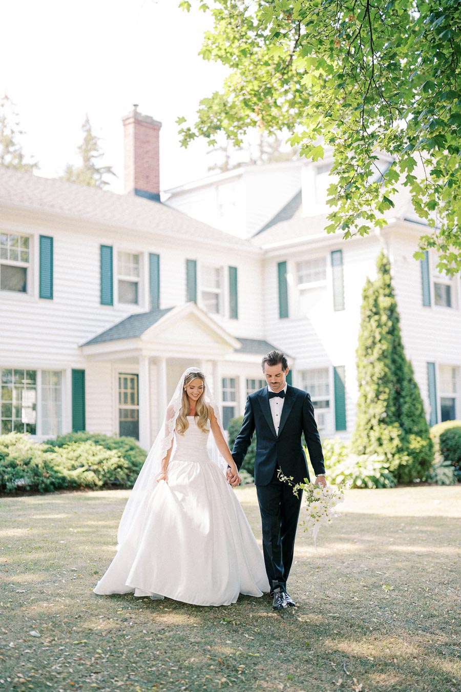 Outdoor wedding couple walking hand in hand, bride in elegant white gown with veil and groom in black tuxedo, in front of classic white house with green shutters under lush green tree.
