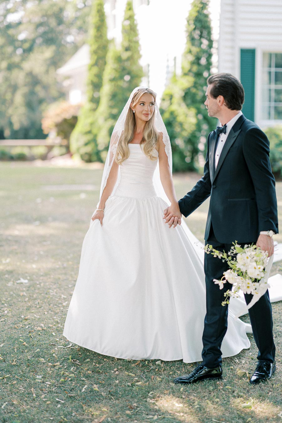 A bride in an elegant white wedding gown and veil holds hands with a groom in a black tuxedo, outdoors in a garden setting with lush greenery.