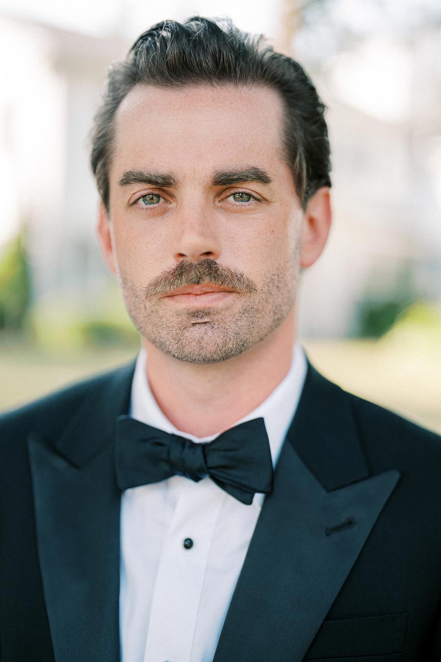 A man with a mustache wearing a formal black tuxedo and bow tie, standing outdoors with a blurred background.