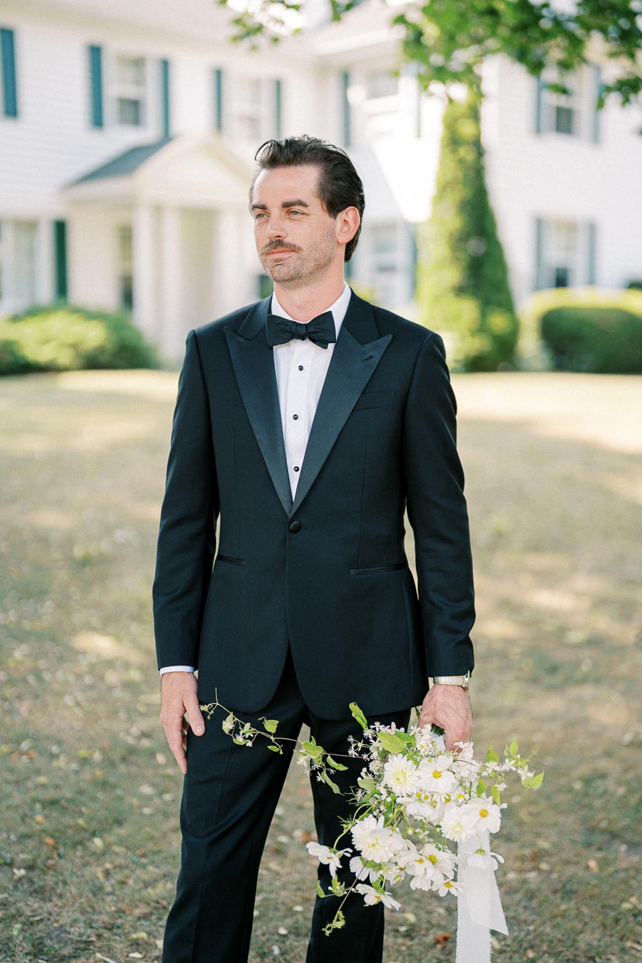 A groom in a black tuxedo holds a bouquet of white flowers, standing outdoors in front of a white house with green shutters.