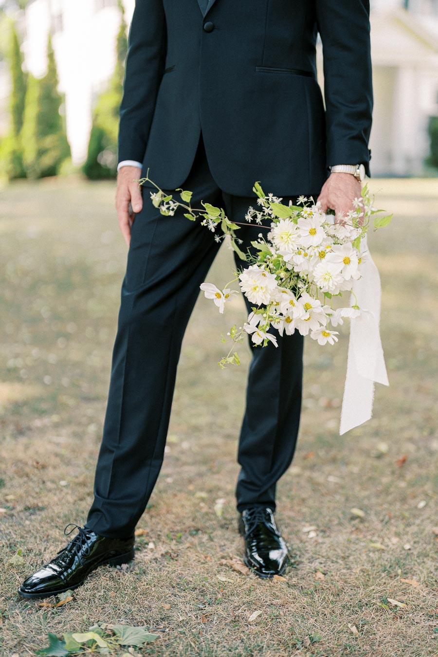 A man in a black suit holds a bouquet of white flowers with greenery, standing on a grassy lawn.