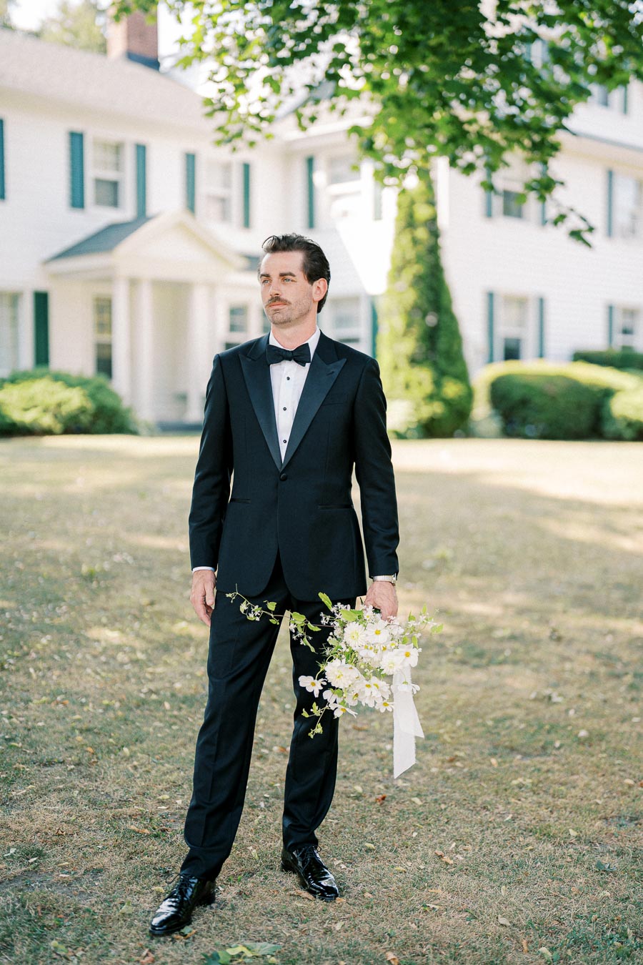 Groom in a black tuxedo holding a bouquet, standing outdoors in front of a white house with green shutters, under lush trees on a sunny day.