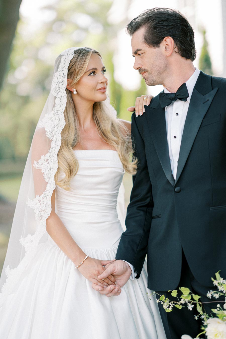 A bride and groom stand lovingly hand in hand, gazing into each other's eyes on their wedding day. The bride wears a white lace dress with a veil, while the groom is in a classic black tuxedo, surrounded by a serene outdoor backdrop.