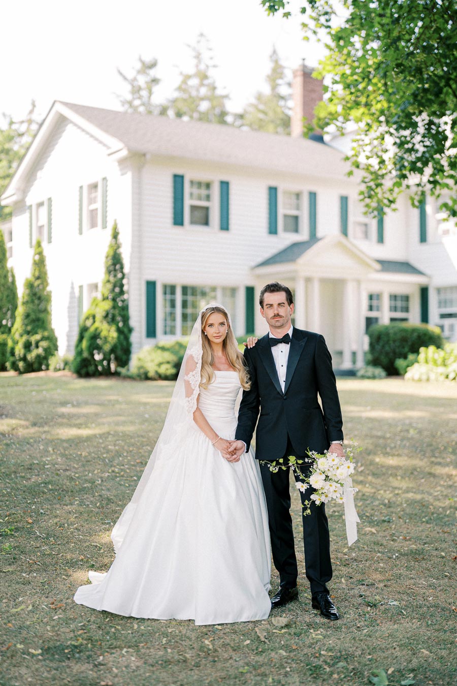 Elegant bride and groom posing in front of a charming white colonial house on their wedding day, the bride in a classic white gown with veil and the groom in a black tuxedo holding a bouquet.