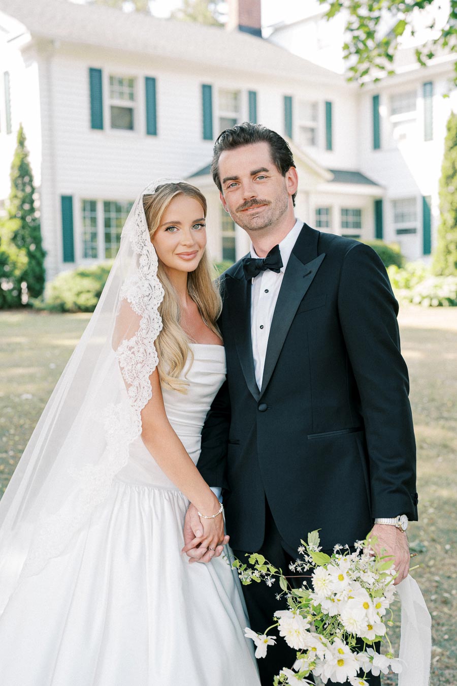 Elegant bride and groom holding hands in front of a classic white house, with the bride wearing a lace-trimmed veil and the groom in a black tuxedo, capturing a timeless wedding moment.