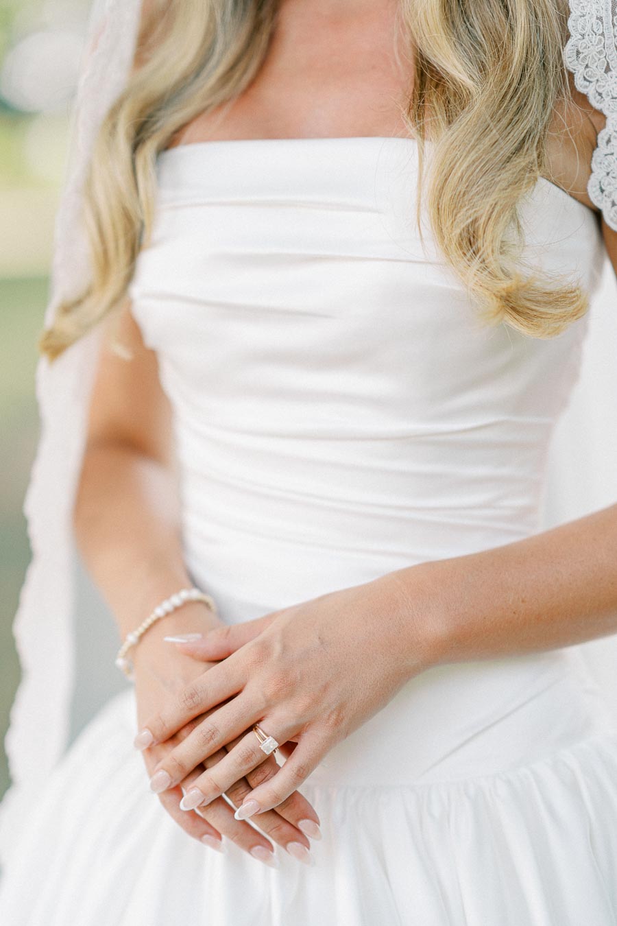 Bride in a white strapless wedding dress with elegant lace details, showcasing her hands adorned with a gold engagement ring and pearl bracelet, creating a romantic bridal look.