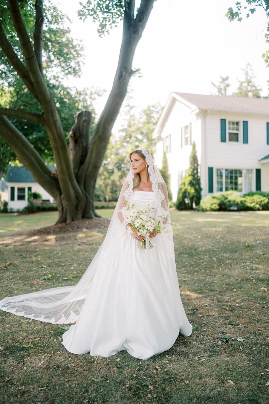 A bride in an elegant white dress and lace veil holding a bouquet of white flowers stands in a sunlit garden near a house, under a large tree.
