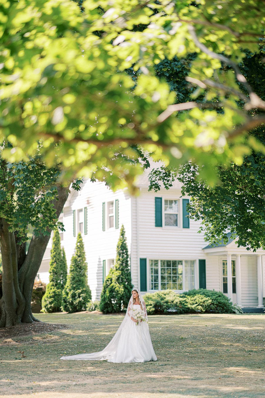 Bride in elegant white gown holding bouquet, standing in front of a charming white house with green shutters, surrounded by lush greenery and large trees.