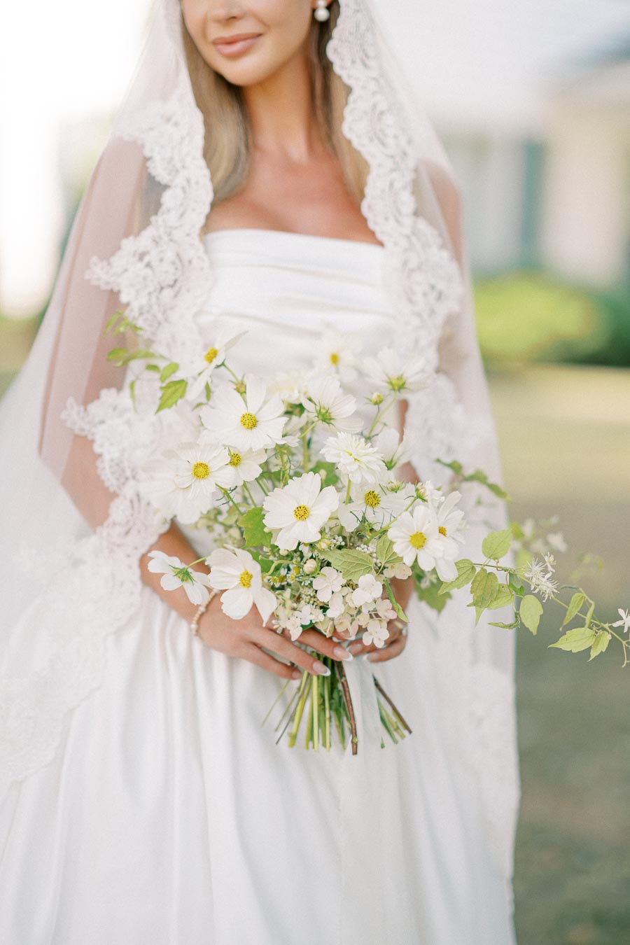 A bride in a white gown and lace veil holding a bouquet of white and green flowers, set against a soft outdoor background.