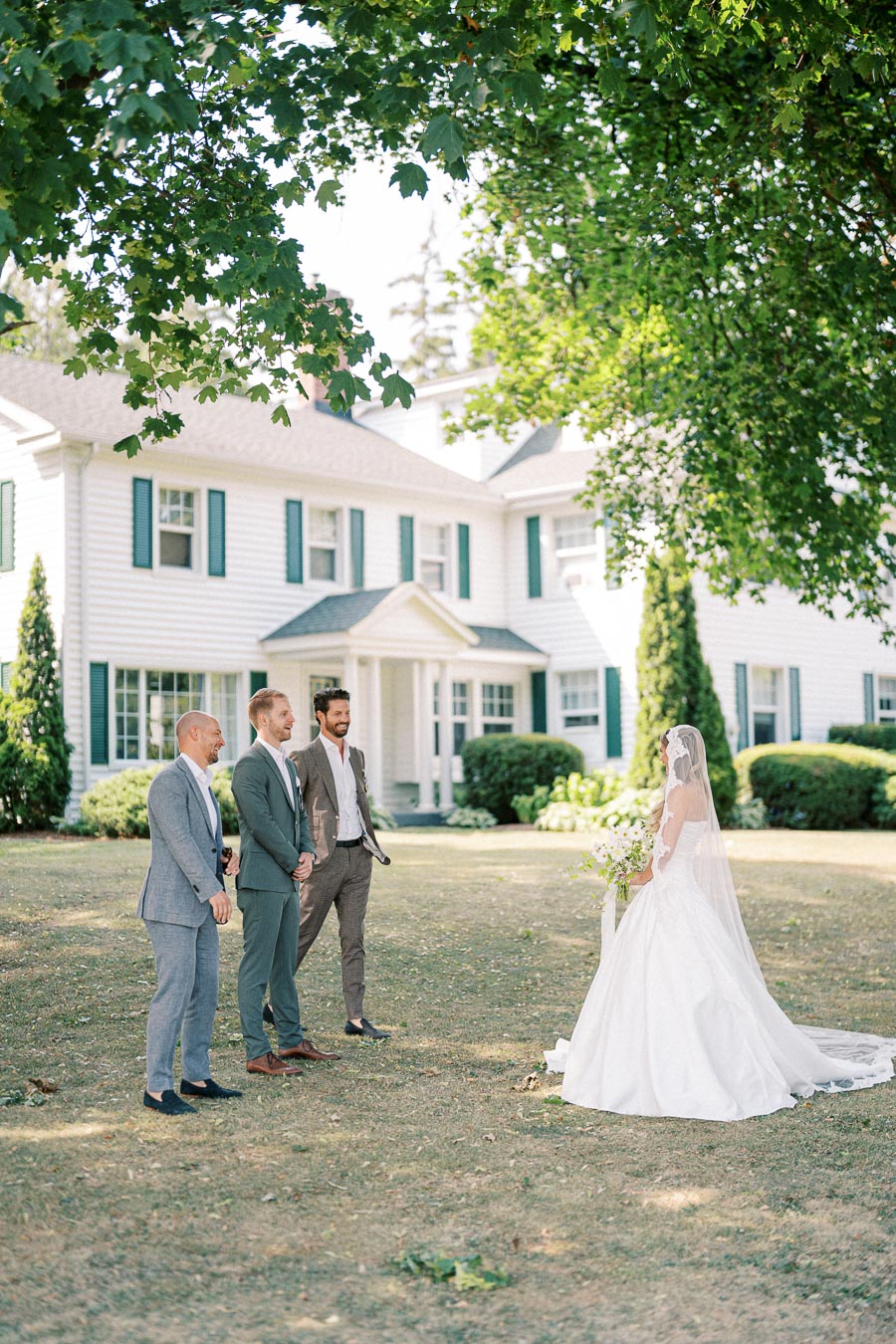 A bride in a white gown and veil standing in a garden surrounded by greenery, with three men in suits smiling near a large white house with green shutters.