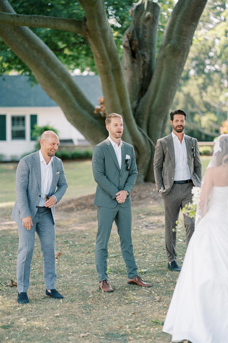 A bride in a white wedding gown walks towards three groomsmen in suits, standing under a large tree during an outdoor wedding ceremony, with a house and greenery in the background.