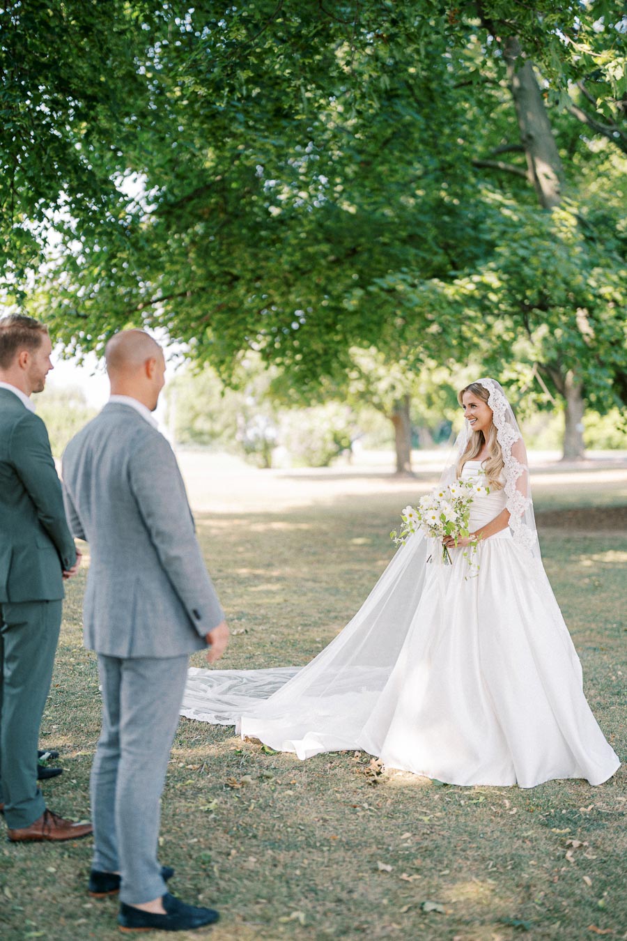 Bride in elegant white gown and lace veil holding a bouquet, standing outdoors under lush green trees during a wedding ceremony, with two groomsmen in suits looking on.