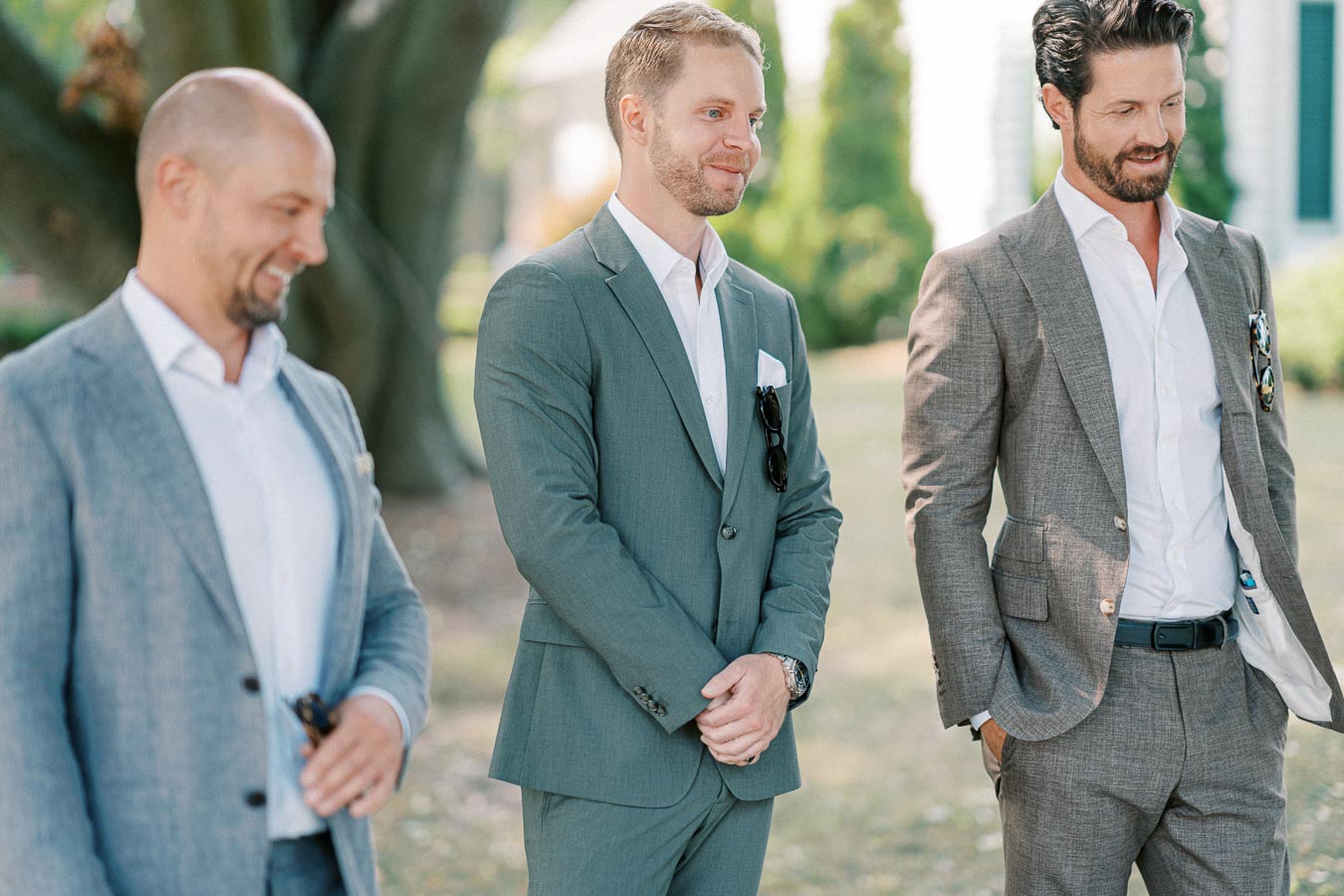Three men in stylish suits standing outdoors with relaxed expressions, possibly at a formal event or gathering, with greenery in the background.