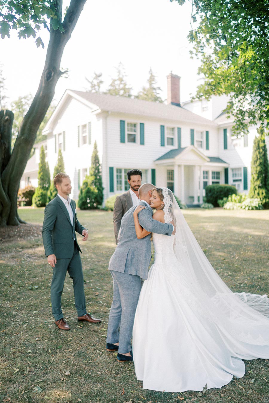 A bride in a white wedding gown and veil embraces a man in a gray suit outside a large white house surrounded by trees, with two other men in suits nearby on a sunny day.