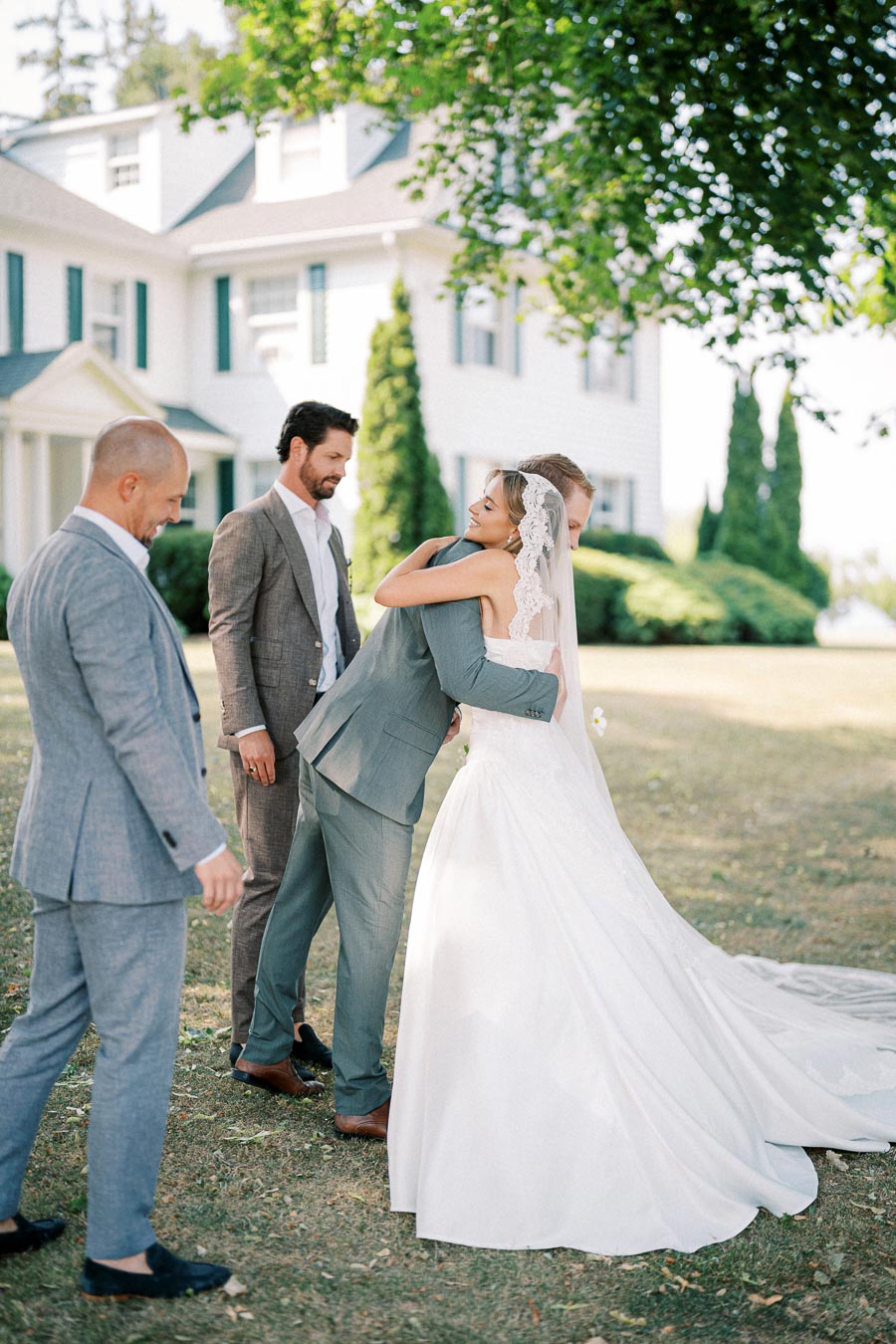 Bride in elegant white gown hugging a groomsman outdoors on a sunny day, with guests in suits nearby, in front of a charming white house with green shutters.