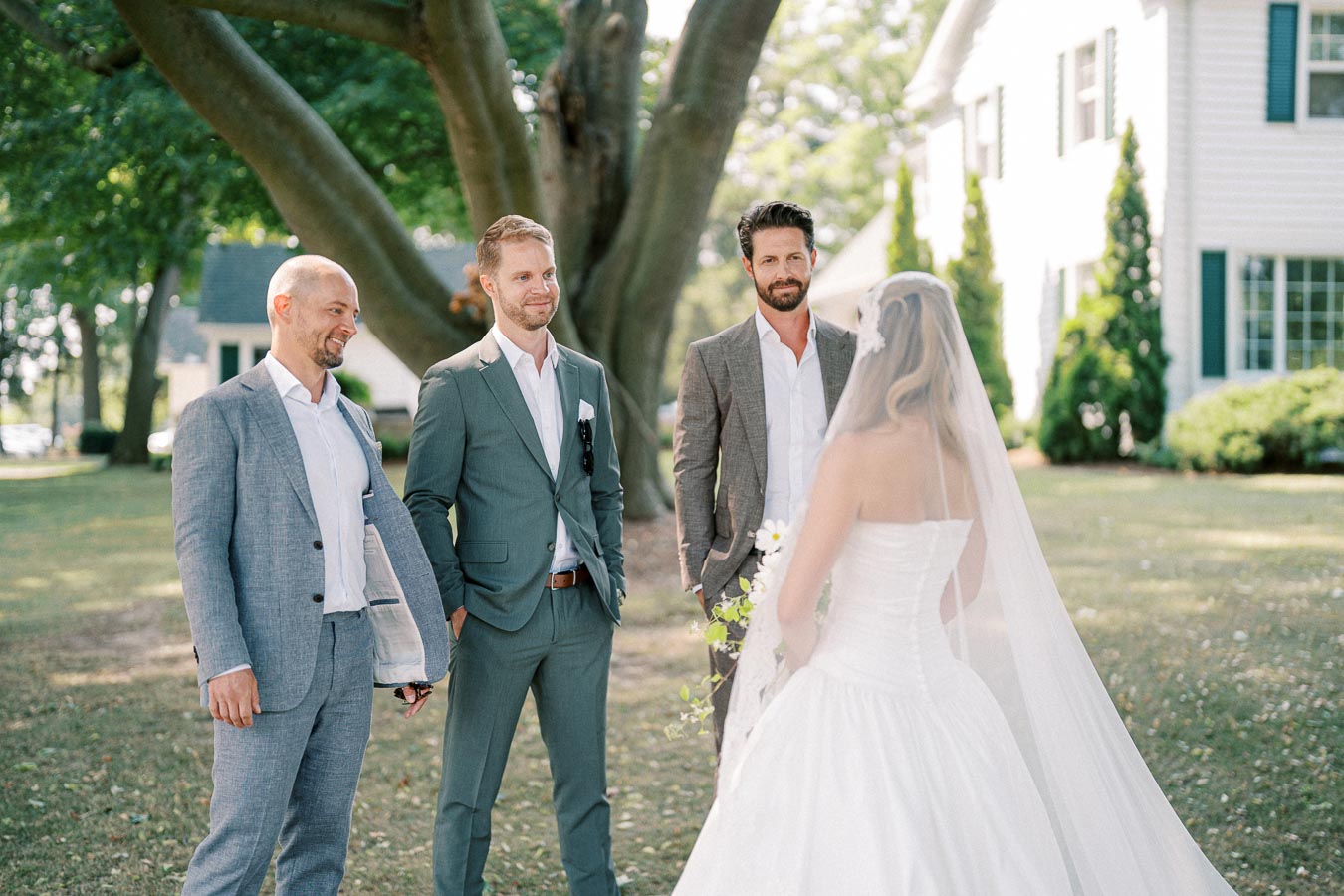 A bride in a white wedding dress with a veil is having a conversation with three groomsmen in suits outdoors, surrounded by trees and a house in the background.