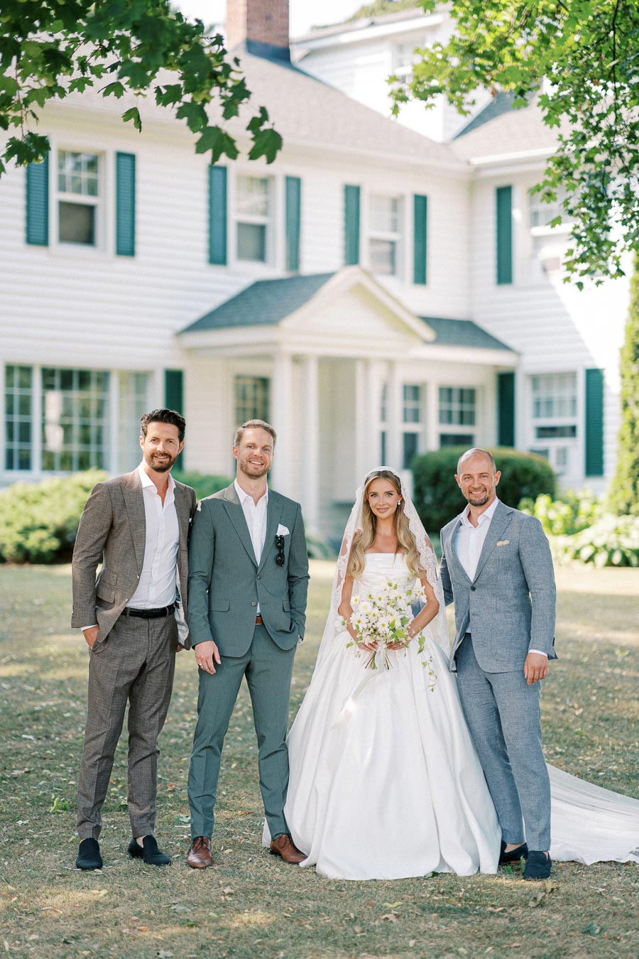 Wedding group portrait in front of a charming white house. The bride stands in an elegant white gown holding a bouquet, surrounded by three groomsmen in stylish suits. The scene is set in a lush garden with a serene and joyful atmosphere.
