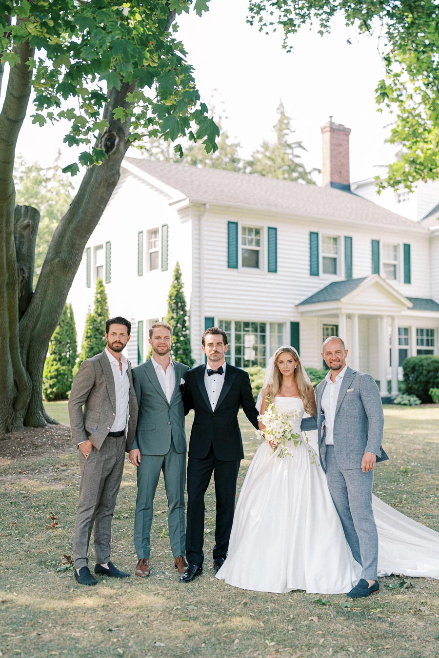 Wedding party posing outdoors in front of a large white house, featuring a bride in a white gown holding a bouquet, flanked by four groomsmen in suits under a large green tree.
