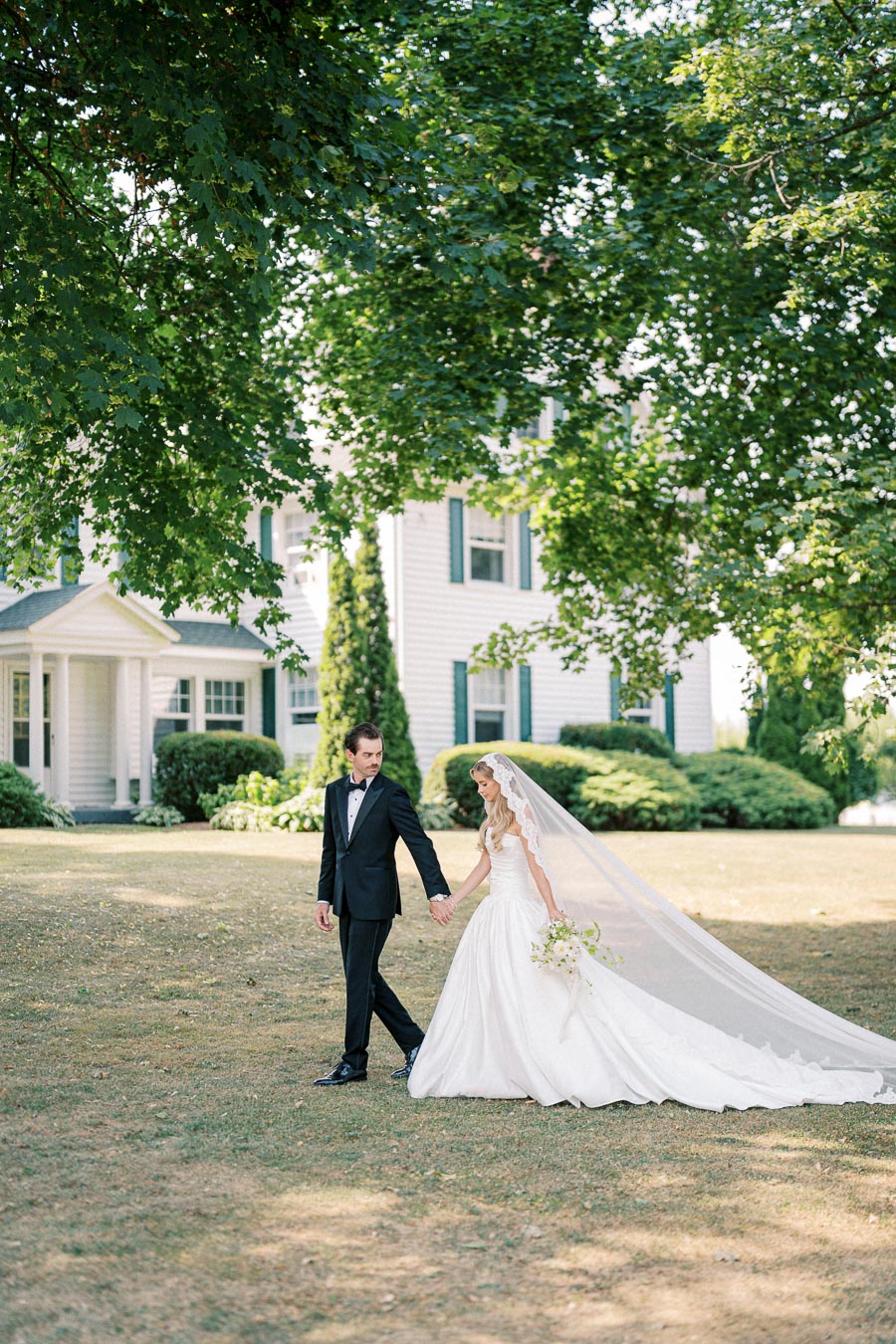 A bride and groom in elegant wedding attire walk hand in hand on a grassy lawn, surrounded by greenery and in front of a charming white house.