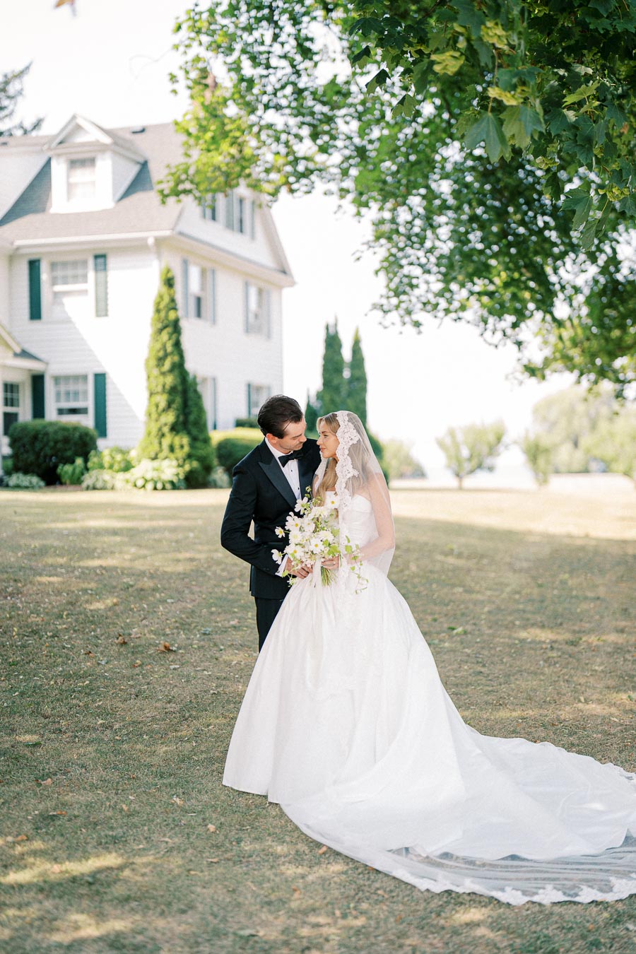 A bride and groom embrace outdoors near a large white house, with the bride in a flowing white dress and holding a bouquet, surrounded by trees and greenery on a clear day.