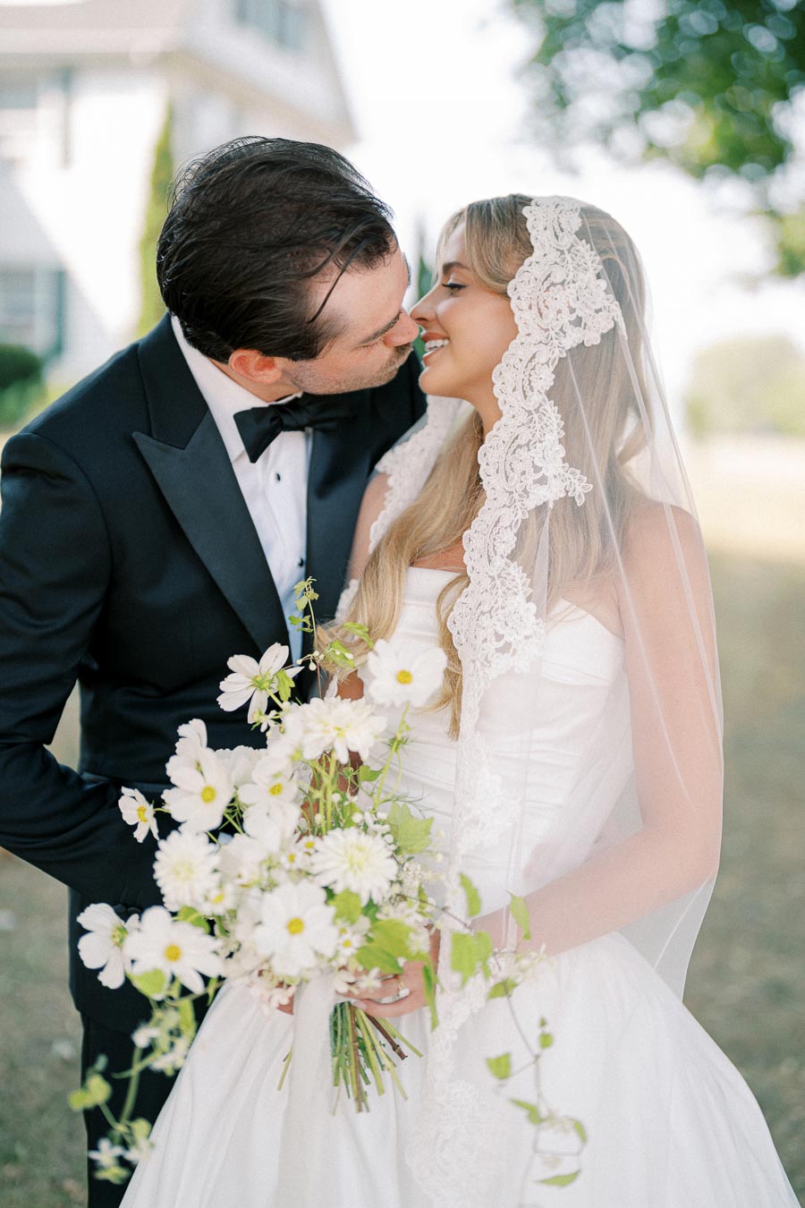 A bride and groom share a romantic kiss outdoors, with the bride holding a bouquet of white flowers. The bride is wearing a lace veil, and the groom is in a black tuxedo.